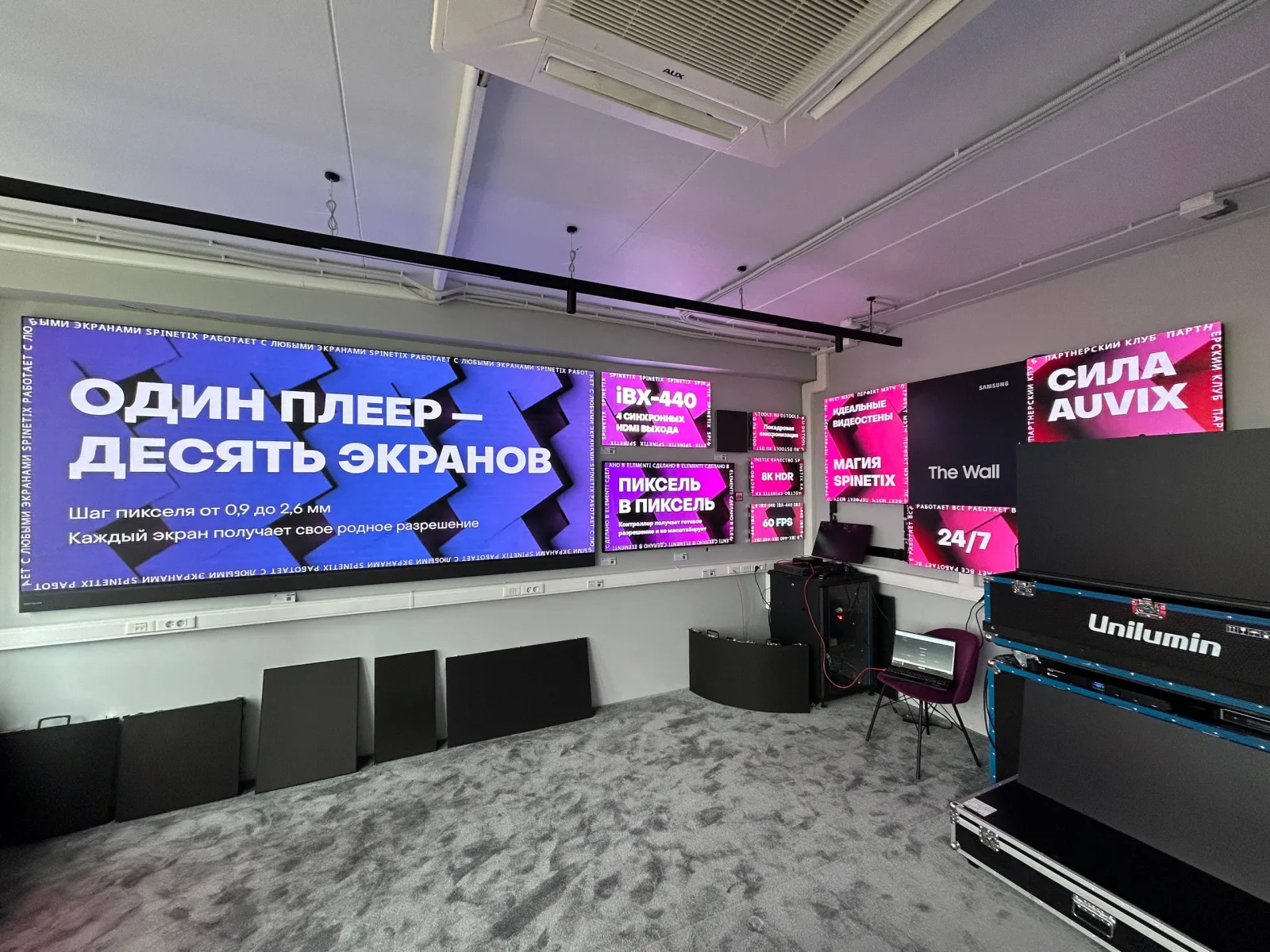 Та же стена в шоуруме AUVIX в малиново-фиолетовой сцене: угловатый геометрический фон проходит через все девять экранов, включая мелкие панели справа.