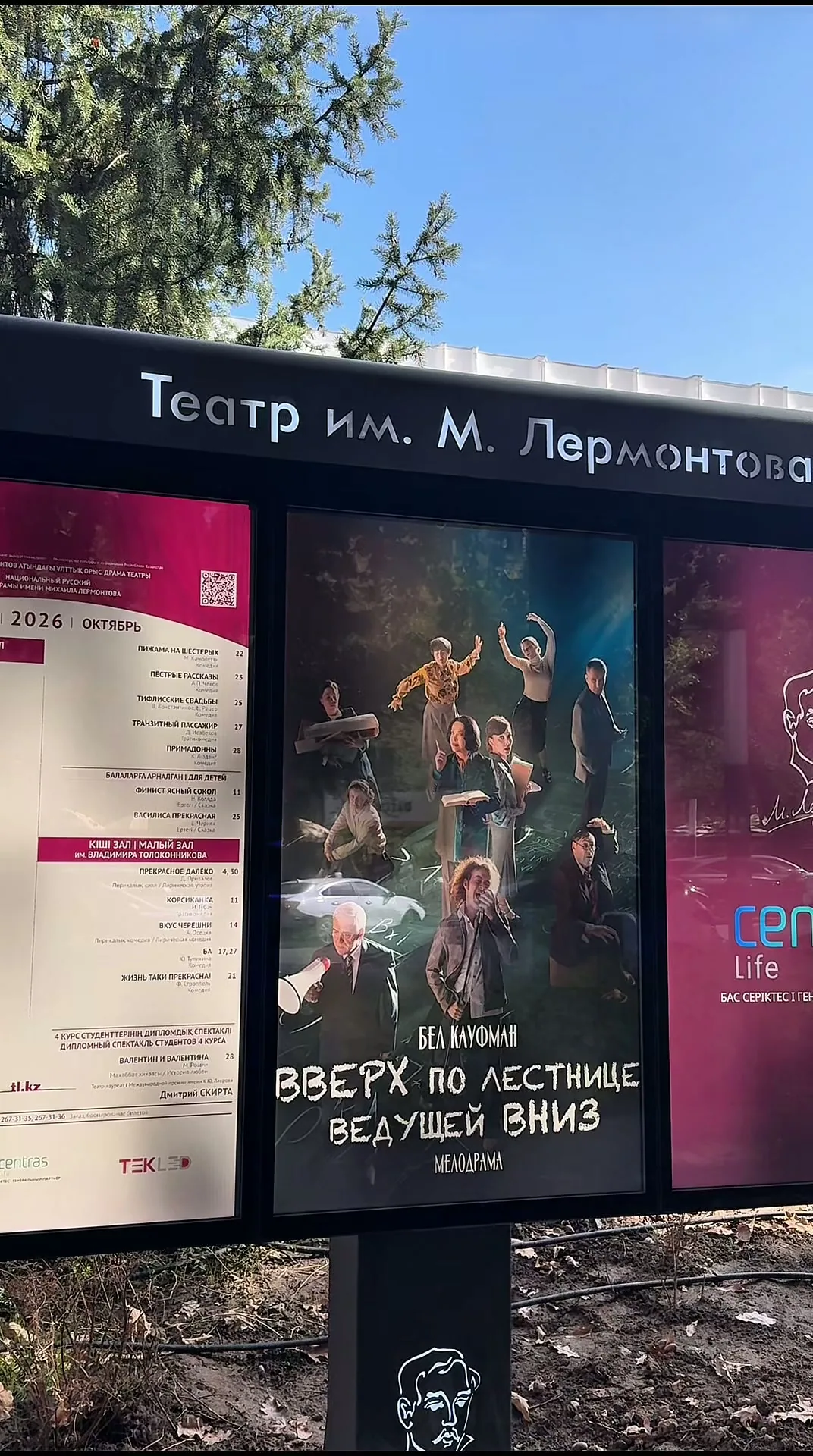 Уличный афишный стенд театра имени Лермонтова: слева печатное расписание на октябрь 2026 года, в центре афиша спектакля «Вверх по лестнице, ведущей вниз», справа вертикальный экран с логотипом Centras Life